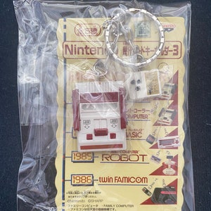 Puede incluir: Un llavero con una réplica en miniatura de la consola Nintendo Family Computer (NES), un mando y un cartucho de juego. El llavero está hecho de metal y plástico y tiene un esquema de color rojo y blanco. El llavero también incluye el texto "Nintendo Family Computer" y "Twin Famicom" junto con los años 1985 y 1986.
