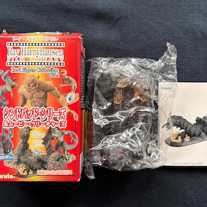 May include: A red box featuring a monster illustration on the front. The box is labelled "Ray Harryhausen Film Library Real Figure Collection Vol. 4". The box also displays the text "Sindbad Series & Movie Creatures". The box is positioned next to a clear plastic bag containing a dark brown figure. A white card with the word "Furuta" is also visible.