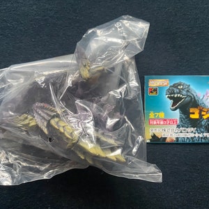 Peut inclure: Un sac en plastique contenant un jouet Godzilla violet et jaune. Le sac est à côté d'une carte avec le texte "Godzilla 7" et une image de Godzilla.