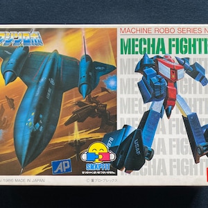 Może przedstawiać: Pudełko z zestawem modeli vintage przedstawiające niebieskiego i czerwonego robota, czarny odrzutowiec i tekst "MACHINE ROBO SERIES No.6 MECHA FIGHTER". Pudełko jest białe z niebieskimi i czerwonymi akcentami. Robot trzyma broń i ma niebiesko-czerwoną kolorystykę. Odrzutowiec to czarny SR-71 Blackbird.