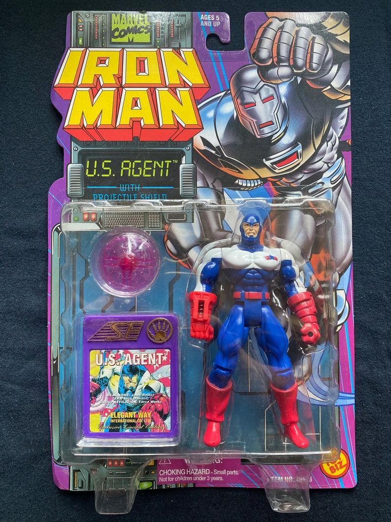 K&ouml;nnte beinhalten: Eine Plastik-Actionfigur von U.S. Agent aus Marvel Comics. Die Figur tr&auml;gt ein rot-wei&szlig;-blaues Kost&uuml;m mit einem rot-wei&szlig;en Schild am linken Arm. Die Figur ist in einer Plastik-Blisterverpackung mit einem violetten Projektilschild verpackt.