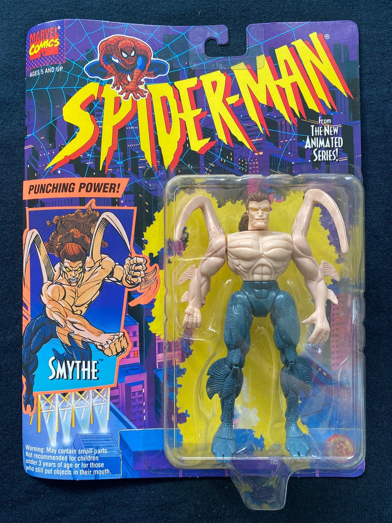 Puede incluir: Una figura de acci&oacute;n de pl&aacute;stico del personaje Smythe de la serie animada de Spider-Man. La figura est&aacute; en un paquete azul y amarillo con las palabras "Spider-Man" y "Punching Power!" en &eacute;l. La figura lleva un atuendo azul y gris con un cuerpo marr&oacute;n claro. La figura tiene un par de alas en la espalda.