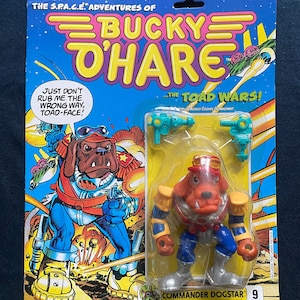 Könnte beinhalten: Eine Plastik-Actionfigur von Commander Dogstar aus der S.P.A.C.E. Adventures of Bucky O'Hare Spielzeuglinie. Die Figur ist rot, blau und silber mit einem gelben Stern auf seinem Hut. Die Figur hält eine Laserpistole in ihrer rechten Hand. Die Verpackung ist blau und gelb mit einer Comic-Illustration von Commander Dogstar und dem Text "The Toad Wars!"