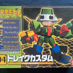 May include: A Custom Robo action figure box featuring a green and blue robot with red and yellow accents. The box has Japanese text and the number "04". The box also displays the Custom Robo logo and other robot designs.