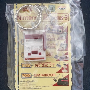 May include: A keychain featuring a miniature replica of the Nintendo Family Computer (NES) console. The keychain is made of plastic and has a red and white color scheme. The keychain also includes a small metal ring for attaching to keys or a bag. The packaging features images of the NES, the Family Computer Robot, and the Twin Famicom, along with the text "Licensed by Nintendo" and "Banpresto".