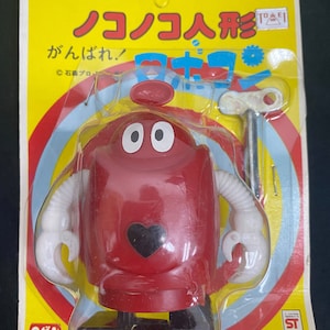 Puede incluir: Un robot de juguete rojo de cuerda con un corazón negro en el pecho y brazos blancos. El robot está empaquetado en una caja de plástico transparente con fondo amarillo y texto japonés que dice "Nokonoko Robot" y "Gankonbare!" El paquete también incluye una llave de cuerda plateada.