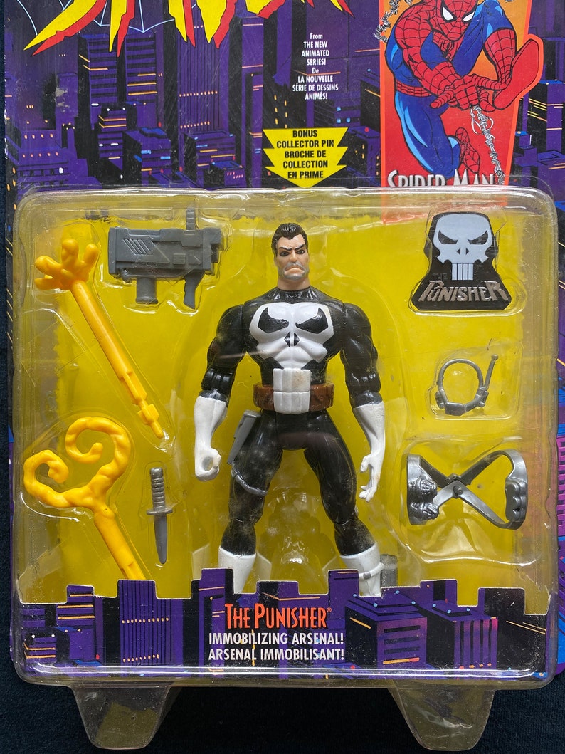 May include: A plastic action figure of The Punisher from the animated series Spider-Man. The Punisher is wearing his signature black and white costume with a skull emblem on his chest. The figure comes with a variety of accessories, including a gun, a whip, and a grappling hook. The packaging features a background of city buildings and the text "The Punisher" and "Immobilizing Arsenal!"