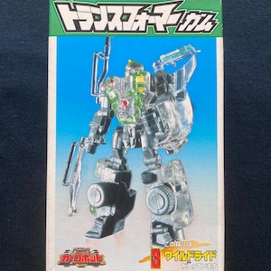 Könnte beinhalten: Ein Spielzeug in einer Schachtel mit einer durchsichtigen und grauen Roboterfigur mit grünen und gelben Akzenten. Die Schachtel hat japanischen Text und einen grün-blauen Hintergrund. Das Spielzeug ist eine Transformers Wildride Figur.