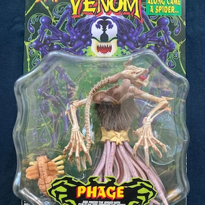 May include: A plastic package containing a toy action figure of Phage, a character from the Spider-Man Venom comic book series. The figure is beige and has a long, thin body with multiple legs. The figure is posed with its arms outstretched and its legs spread apart. The package is green and black and features the Spider-Man Venom logo. The text on the package reads "Along Came A Spider..." and "Phage TM".