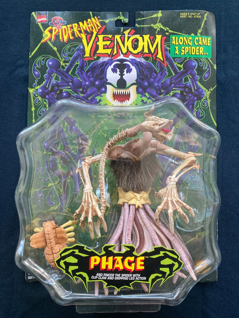 May include: A plastic package containing a toy action figure of Phage, a character from the Spider-Man Venom comic book series. The figure is beige and has a long, thin body with multiple legs. The figure is posed with its arms outstretched and its legs spread apart. The package is green and black and features the Spider-Man Venom logo. The text on the package reads "Along Came A Spider..." and "Phage TM".