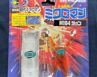 May include: A vintage M104 John Microman action figure on its original packaging. The packaging features Japanese text, a clear plastic case, and a red and orange color scheme. The figure is orange and silver.