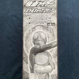 May include: A black and white box featuring a Kamen Rider figure. The box has Japanese text and graphics, including the words "Bottle Cap Stamp". The figure is in a dynamic pose, with a silver helmet and a detailed costume.