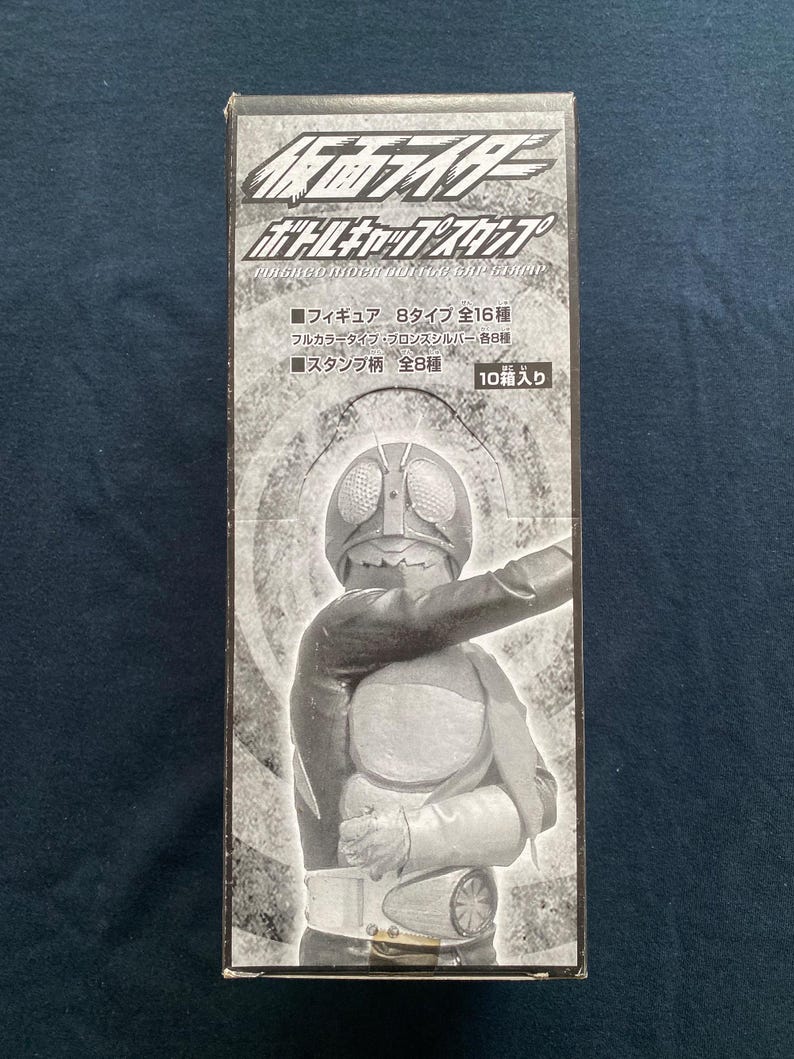 May include: A black and white box featuring a Kamen Rider figure. The box has Japanese text and graphics, including the words "Bottle Cap Stamp". The figure is in a dynamic pose, with a silver helmet and a detailed costume.