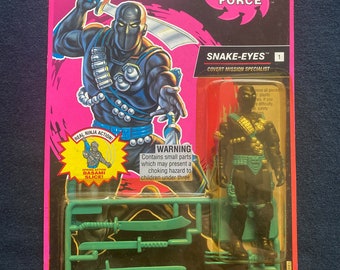 Puede incluir: Una figura de acción vintage de Snake-Eyes de la línea G.I. Joe Ninja Force. La figura está vestida de negro y verde con un cinturón dorado y sostiene una espada. La figura está empaquetada en una caja de cartón rosa y negra con el logotipo de G.I. Joe y las palabras "Ninja Force" impresas en ella. La caja también incluye una advertencia sobre las piezas pequeñas que pueden ser un peligro de asfixia para los niños menores de tres años.