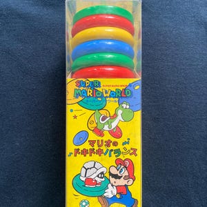 Pode incluir: Um conjunto de brinquedos empilháveis Super Mario World numa caixa de plástico transparente. O conjunto inclui seis anéis coloridos em vermelho, amarelo, azul e verde. A caixa apresenta Mario, Yoshi e outras personagens.