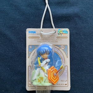 Op de afbeelding: Een klein plastic beeldje van Rei Ayanami uit Neon Genesis Evangelion, gekleed in haar kenmerkende witte pilotenpak en met een boog en pijl in de hand. Het beeldje staat op een voetstuk met het sterrenbeeld Boogschutter en een schild. De verpakking is goud en wit met de tekst "Neon Genesis Evangelion Star and Constellation Mini Display Figure" en "GAINAX".