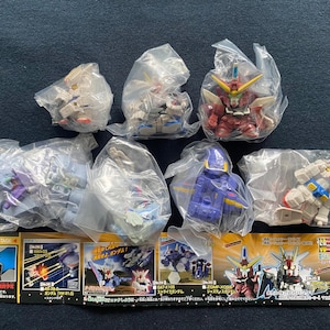 May include: A collection of seven plastic toy robots in various colours, including red, blue, white, and grey. The robots are packaged in clear plastic bags and are displayed on a white surface. The packaging includes the text "THE NEXT STAGE" and "2003年5月発".