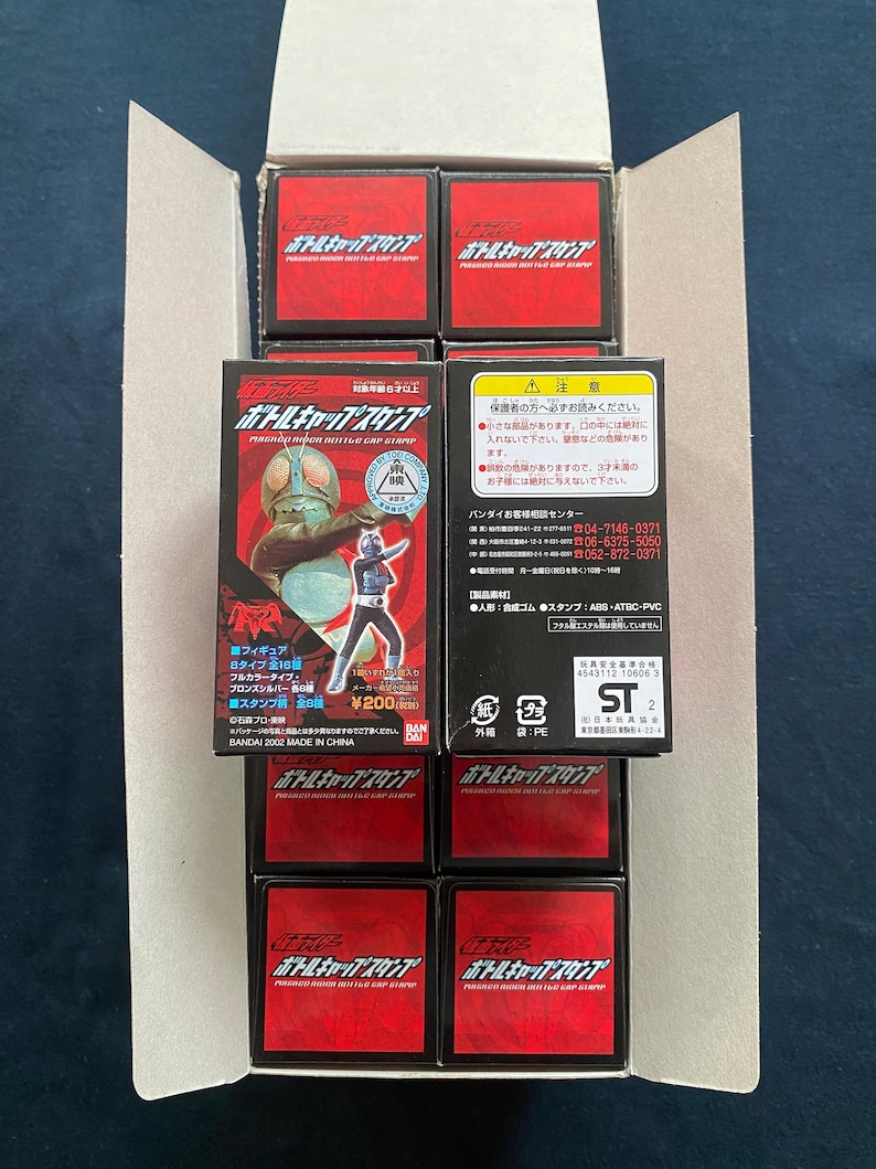 May include: An open cardboard box filled with several red and black toy boxes. Each box features Japanese text and graphics. Some boxes display images of characters. The boxes are stacked inside the cardboard container.