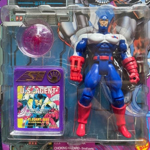 K&ouml;nnte beinhalten: Eine Plastik-Actionfigur von U.S. Agent, einem Superhelden aus Marvel Comics. Die Figur tr&auml;gt ein blaues, wei&szlig;es und rotes Kost&uuml;m mit einem roten Helm. Die Figur h&auml;lt eine rot-schwarze Waffe in ihrer rechten Hand. Die Figur ist in einer Plastik-Blisterverpackung mit violettem Hintergrund verpackt. Die Verpackung enth&auml;lt ein kleines Comic-Heft mit U.S. Agent.