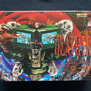 May include: A box for a 1:8 scale model of a robot with a green, black, and red color scheme. The box features the text "Deathtron" in red and white letters. The robot has a large head with multiple screens and a claw-like hand.