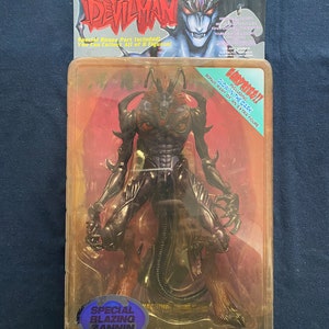 Könnte beinhalten: Eine schwarze und rote Actionfigur eines Dämonencharakters aus der Anime-Serie Devilman in einer Plastikverpackung. Die Figur ist mit ausgestreckten Armen posiert und hat eine Leucht-im-Dunkeln-Funktion. Die Verpackung enthält ein Bonusteil einer zusätzlichen Figur. Der Text auf der Verpackung lautet "Devilman", "Zannin", "Special Bonus Part Included! You Can Collect All of 8 Figures!" und "Glow in the Dark! Including Bonus Part of an Extra Figure!"