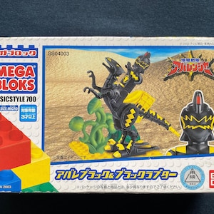 Pode incluir: Uma caixa para Mega Bloks Basicstyle 700, com um brinquedo de dinossauro preto e amarelo com uma planta verde. A caixa é etiquetada "Mega Bloks Basicstyle 700" e "Alpa Black & Black Raptor".