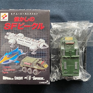Puede incluir: Una caja para un conjunto de juguetes vintage que incluye una nave espacial, un vehículo azul y amarillo y un vehículo militar verde. La caja está etiquetada con "懐かしの SF ビークル", que se traduce como "Vehículos SF nostálgicos".