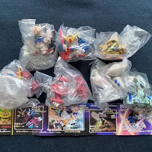 May include: A selection of small, colourful robot figurines in clear plastic packaging. The robots are various colours, including red, blue, yellow, and white. Some have swords or other accessories. The image also shows small, colourful cards with Japanese text.
