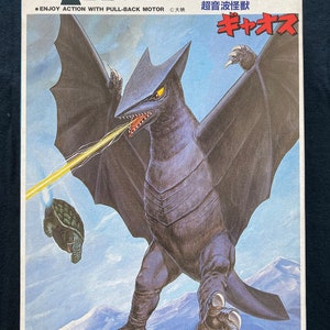 Può includere: Una scatola per un kit di modellismo in plastica del kaiju Gygaos del franchise di Godzilla. La scatola presenta un'illustrazione in nero e blu di Gygaos con un raggio di energia giallo che spara dalla sua bocca. Il testo sulla scatola recita "Scala 1/450 Mostro", "Corri! Serie di mostri a molla", "Mostro ultrasonico", e "Gygaos".