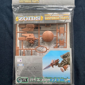 Op de afbeelding: Een verzegelde plastic verpakking met een bruine Zoids Customize Parts modelbouwkit. De verpakking heeft het TOMY-logo en Japanse tekst. De kit bevat diverse bruine plastic onderdelen, waaronder een schild en wapenonderdelen.