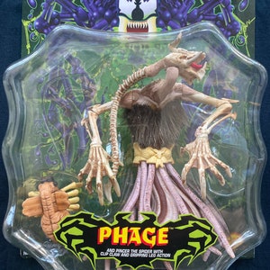 May include: A plastic package containing a Phage action figure from the Venom toy line. The figure is a multi-legged, spider-like creature with a brown body and purple legs. The packaging has a green background with a black and purple Venom character graphic. The text on the packaging reads "Phage" and "And Pince the Spider with Clip Claw and Gripping Leg Action."