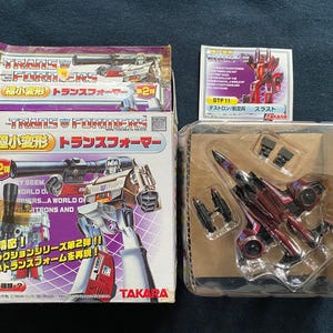 Peut inclure: Deux jouets Transformers, l'un un robot rouge et noir et l'autre un robot gris et argent, sont présentés dans leur emballage. L'emballage est rouge, blanc et bleu avec du texte japonais. Le texte sur l'emballage indique "Transformers" et "Mini-Transformers".
