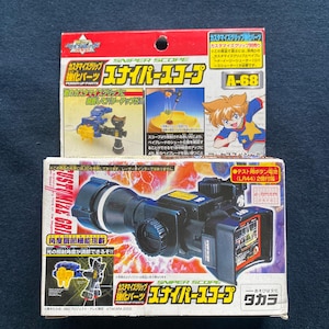 Pode incluir: Duas caixas de brinquedos com a etiqueta "Sniper Scope" com texto e ilustrações japonesas. A caixa superior é vermelha e amarela, com uma personagem de desenho animado. A caixa inferior é preta e branca, mostrando uma mira com funções ajustáveis.