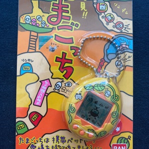 May include: A yellow and orange Tamagotchi keychain with a green vine design. The screen shows a pixelated image of a creature. The packaging features a cartoon illustration of a forest with a path leading to a cave. The text on the packaging reads "Tamagotchi" and "BAN DAI".