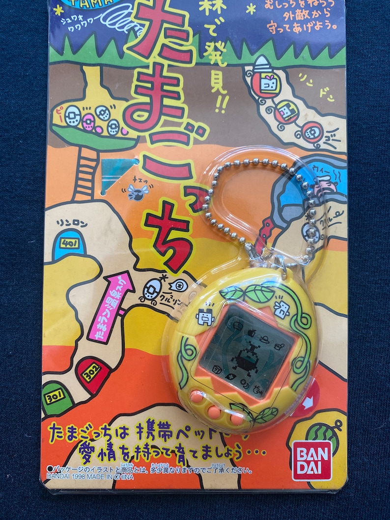 May include: A yellow and orange Tamagotchi keychain with a green vine design. The screen shows a pixelated image of a creature. The packaging features a cartoon illustration of a forest with a path leading to a cave. The text on the packaging reads "Tamagotchi" and "BAN DAI".