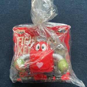 Puede incluir: Un llavero de robot rojo con un corazón en el pecho y manos verdes. El llavero está empaquetado en una bolsa de plástico con una tarjeta roja y blanca que dice "FIGHTER KEY".