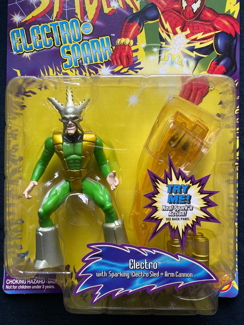May include: A green and gold action figure of Electro from the Spider-Man comics. The figure is holding a yellow and gold arm cannon. The packaging features a yellow background with the text "Electro Spark" and "Try Me! Real Spark'n Action! See Back Panel".