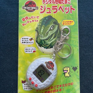 Könnte beinhalten: Ein weißer digitaler Haustier-Schlüsselanhänger in Form eines Dinosauriereis mit einem roten Herzen und einem gelben Dinosaurier auf dem Bildschirm. Die Verpackung zeigt einen grünen Dinosaurier, der aus einem Ei schlüpft, und den Text "The Lost World: Jurassic Park" und "Digital Dinosaur Egg: Jurassic Pet".