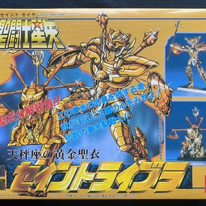 以下が含まれることがあります： 聖闘士星矢のキャラクターが描かれた金色の箱。キャラクターは金色の鎧を着ており、剣を持っている。箱には「聖闘士星矢」と「天秤座」の文字も書かれている。