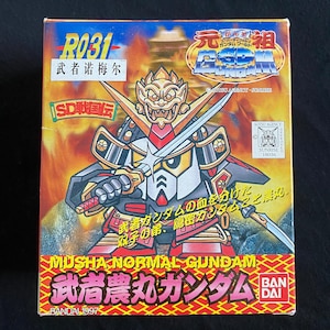 K&ouml;nnte beinhalten: Eine farbenfrohe Box mit einer Cartoon-Illustration eines Samurai-Roboters, der zwei Schwerter h&auml;lt. Die Box hat japanischen Text und die englischen Worte "MUSHA NORMAL GUNDAM". Die Box ist mit "BANDAI 1997" beschriftet.