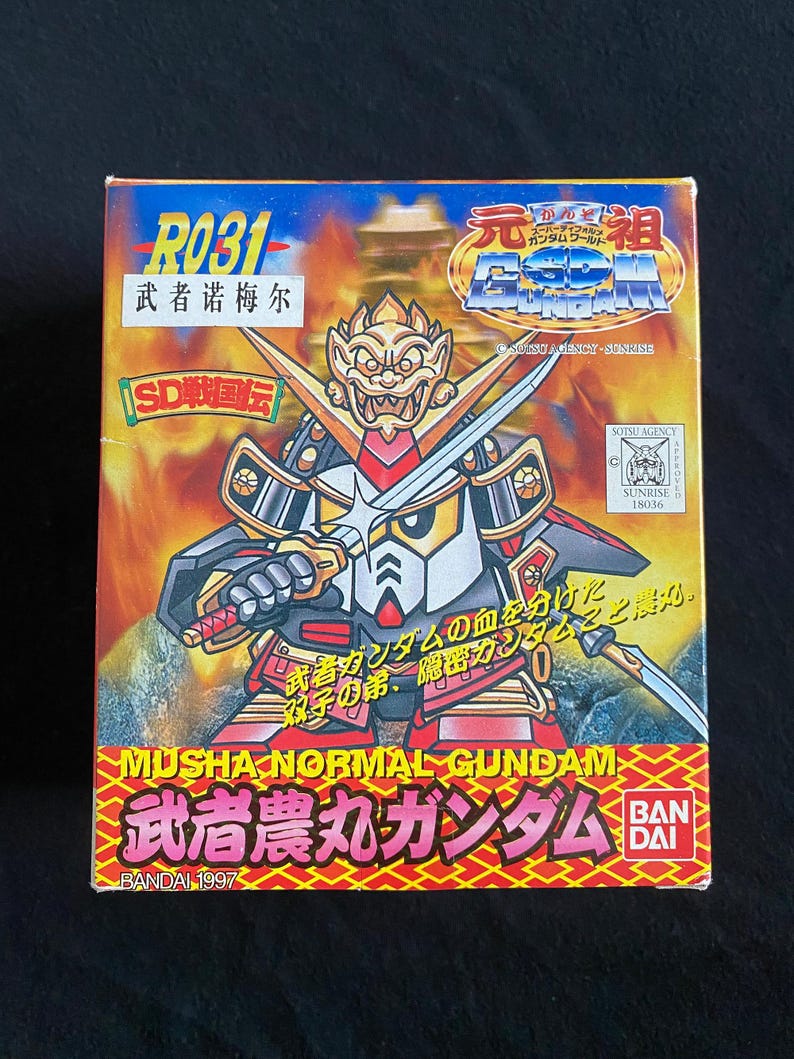 K&ouml;nnte beinhalten: Eine farbenfrohe Box mit einer Cartoon-Illustration eines Samurai-Roboters, der zwei Schwerter h&auml;lt. Die Box hat japanischen Text und die englischen Worte "MUSHA NORMAL GUNDAM". Die Box ist mit "BANDAI 1997" beschriftet.