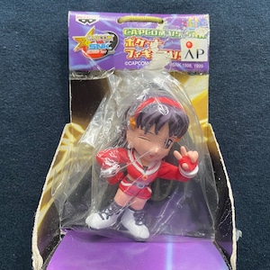 May include: A red and white figure of a female character from the video game Capcom vs. SNK. The figure is holding a peace sign with her right hand. The figure is packaged in a clear plastic box with a yellow and black background. The box has the text "CAPCOM VS. SNK" and "CAPCOM" on it.