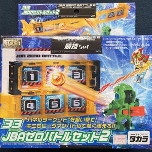 Puede incluir: Una caja amarilla y negra con un juguete dentro. La caja tiene el texto "JBA Zero Battle Set 2" en ella. El juguete es un robot verde con una franja roja y blanca en la espalda. La caja también tiene una imagen de un juguete amarillo y negro.