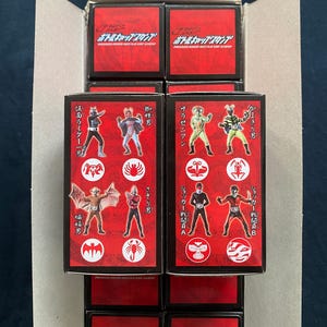 May include: Stacked boxes with red and black packaging. Each box features illustrations of various characters in action poses. The boxes are labeled with Japanese text and character names. The overall theme is action figures or collectibles.