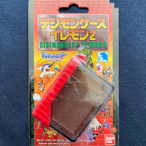 May include: A red and clear plastic case for a Digimon toy. The case has a Digimon logo and the text "Digimon Case Demon 2" on the front. The case is packaged in a red and white cardboard box with the text "Digimon Case Demon 2" on the front.