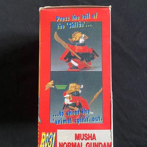 K&ouml;nnte beinhalten: Rote und wei&szlig;e Box mit einer Spielzeug-Samurai-Figur. Der Text lautet "Press the tail of the 'Shii'" und "...to shoot the 'animal spirit' out." Das Spielzeug ist mit "MUSHA NORMAL GUNDAM" beschriftet.