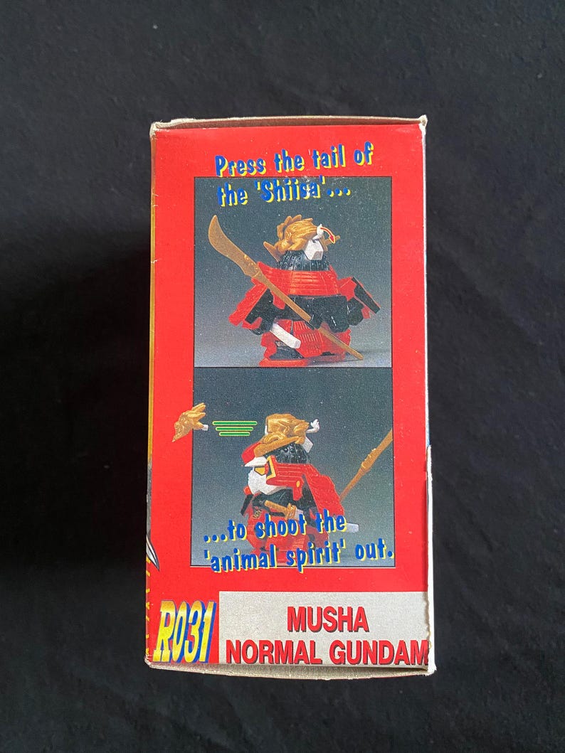 K&ouml;nnte beinhalten: Rote und wei&szlig;e Box mit einer Spielzeug-Samurai-Figur. Der Text lautet "Press the tail of the 'Shii'" und "...to shoot the 'animal spirit' out." Das Spielzeug ist mit "MUSHA NORMAL GUNDAM" beschriftet.
