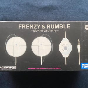Peut inclure: Boîte noire avec le texte "FRENZY & RUMBLE - playing earphone-" et le logo Transformers. La boîte présente des designs d'écouteurs blancs et une prise jack audio de 3,5 mm. Le produit est de Takara Tomy.