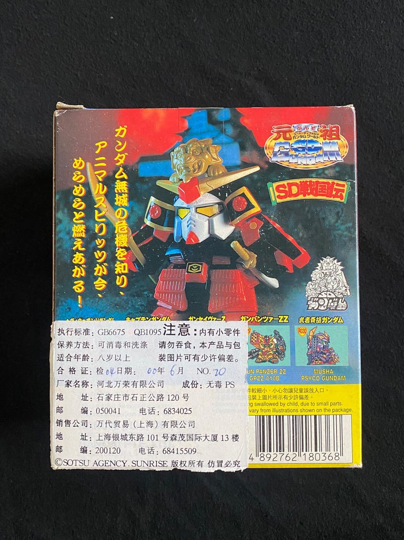 K&ouml;nnte beinhalten: Eine farbenfrohe Box mit einer Gundam-Figur. Die Box enth&auml;lt japanischen Text und Illustrationen des Roboters. Die Figur ist rot, gold und blau. Die Box hat auch Text in Englisch und Chinesisch.