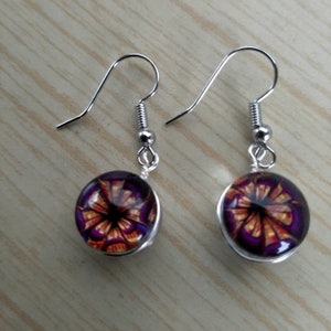 May include: A pair of silver-toned dangle earrings. Each earring features a round, glass-covered charm with a purple, orange, and black floral design. The earrings have a simple hook closure.